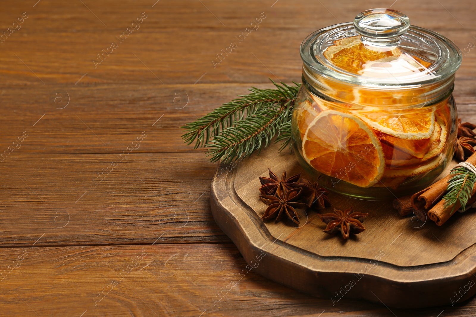 Photo of Glass jar of dry orange slices, anise stars, cinnamon sticks and fir branches on wooden table. Space for text