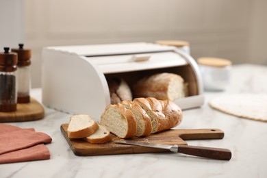 Wooden bread basket with freshly baked loaves and knife on white marble table in kitchen