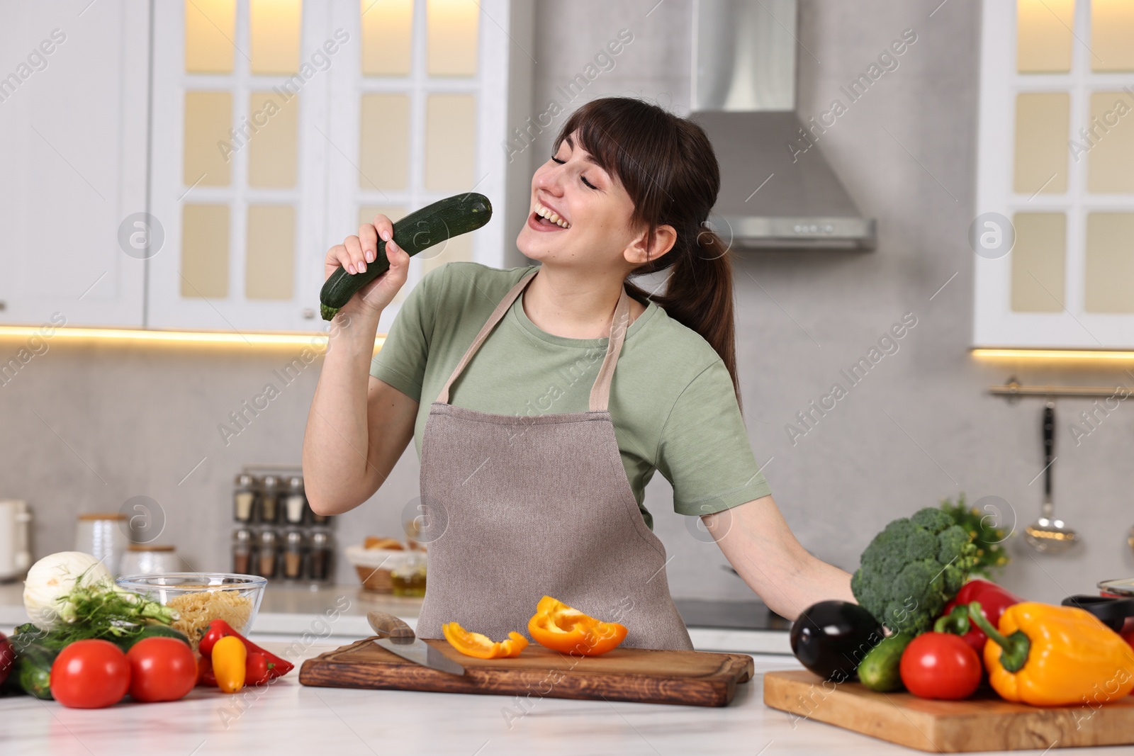 Photo of Happy young housewife with fresh cucumber having fun while cooking at white marble table in kitchen