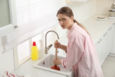 Photo of Upset young woman using plunger to unclog sink drain in kitchen