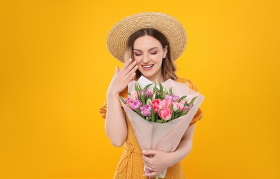 Happy young woman in straw hat holding bouquet of beautiful tulips on yellow background