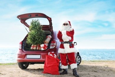 Photo of Authentic Santa Claus near red car with gift boxes and Christmas tree on beach