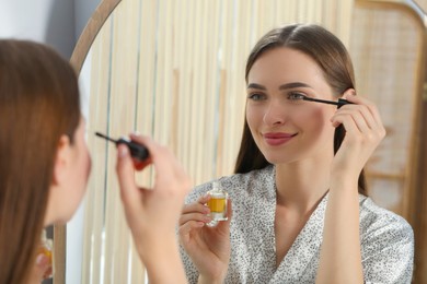 Young woman applying oil on her eyelashes near mirror indoors