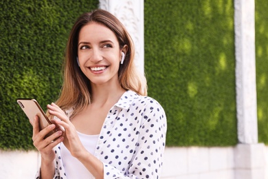 Young woman with wireless headphones and mobile device listening to music near green grass wall. Space for text