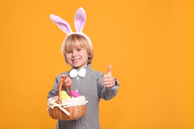 Photo of Happy boy in bunny ears headband holding wicker basket with painted Easter eggs on orange background. Space for text