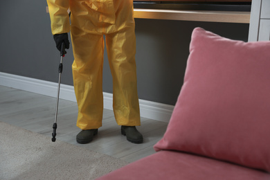 Photo of Pest control worker in protective suit spraying insecticide on floor at home, closeup