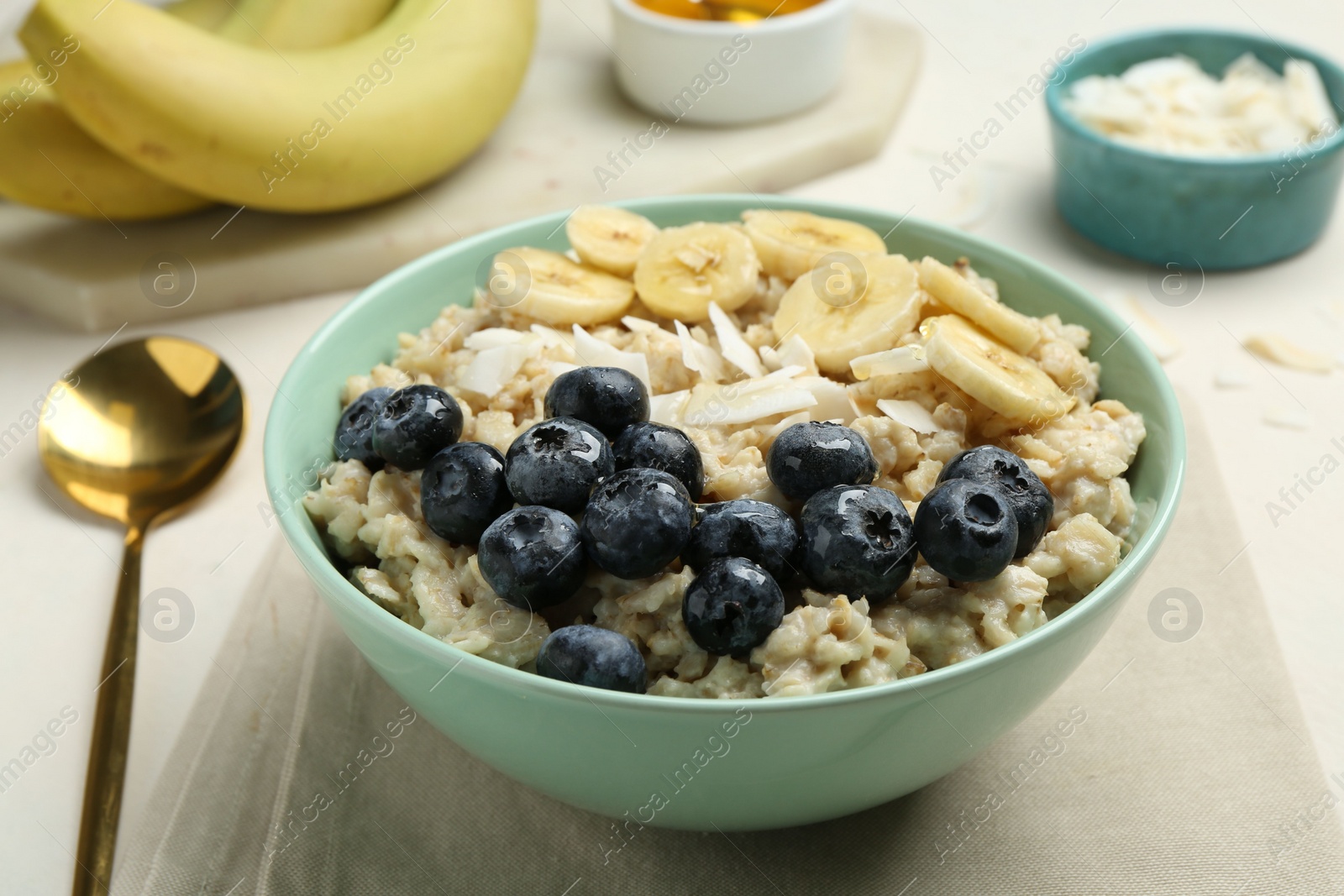 Photo of Tasty oatmeal with banana, blueberries, coconut flakes and honey served in bowl on beige table