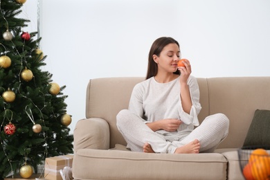 Woman with tangerine sitting on sofa near Christmas tree