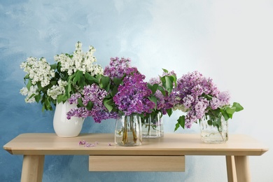 Photo of Vases with beautiful blossoming lilac on table against color background. Spring flowers