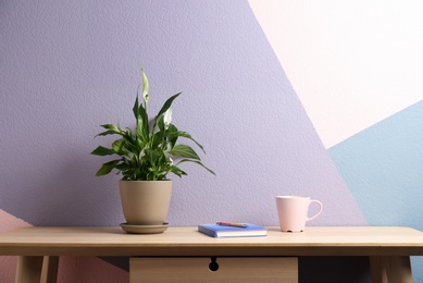 Photo of Potted peace lily plant, cup and notebook on wooden table near color wall. Space for text