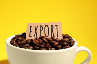 Photo of Coffee beans, white cup and card with word Export on yellow background, closeup