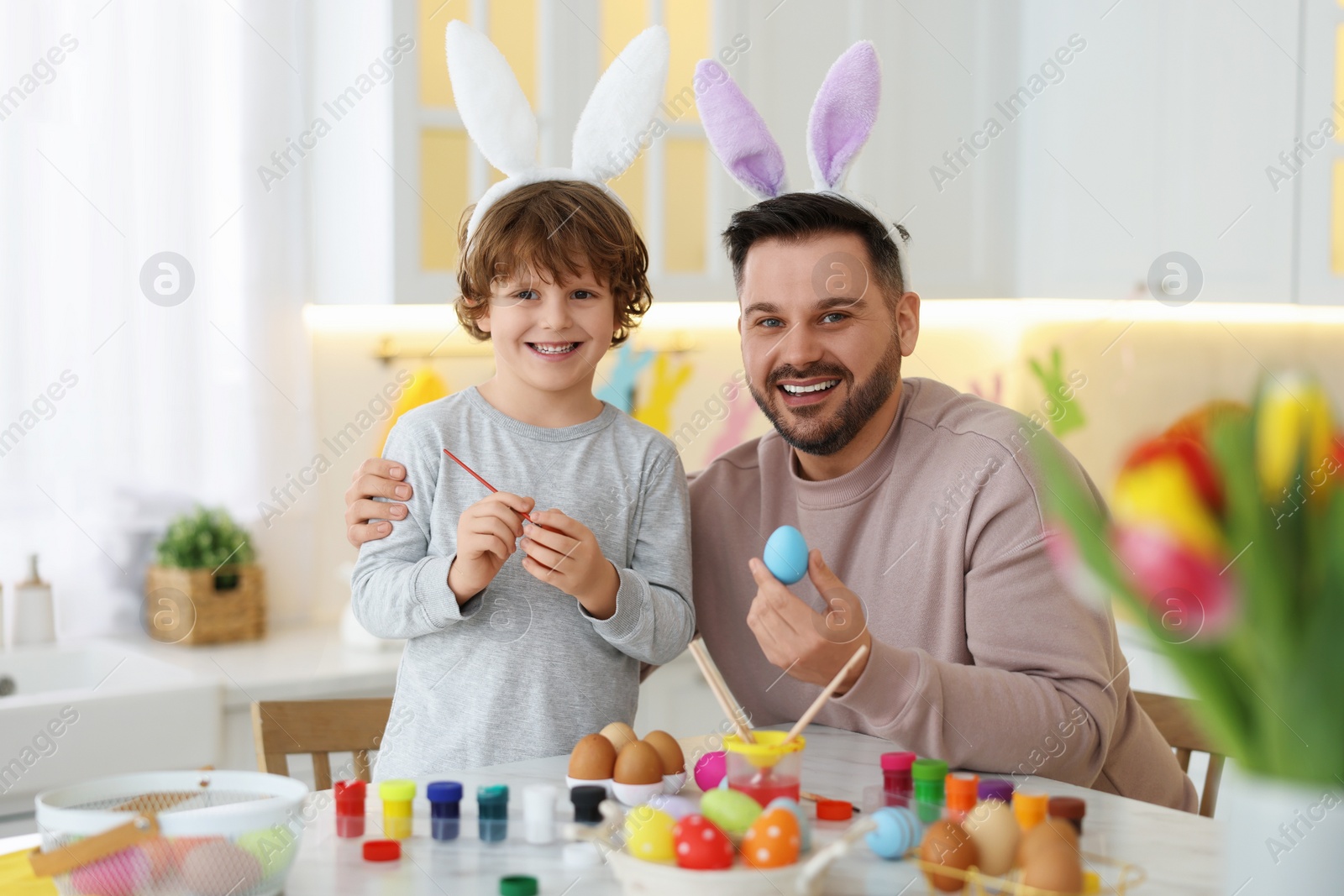 Photo of Easter celebration. Happy father with his little son painting eggs at white marble table in kitchen