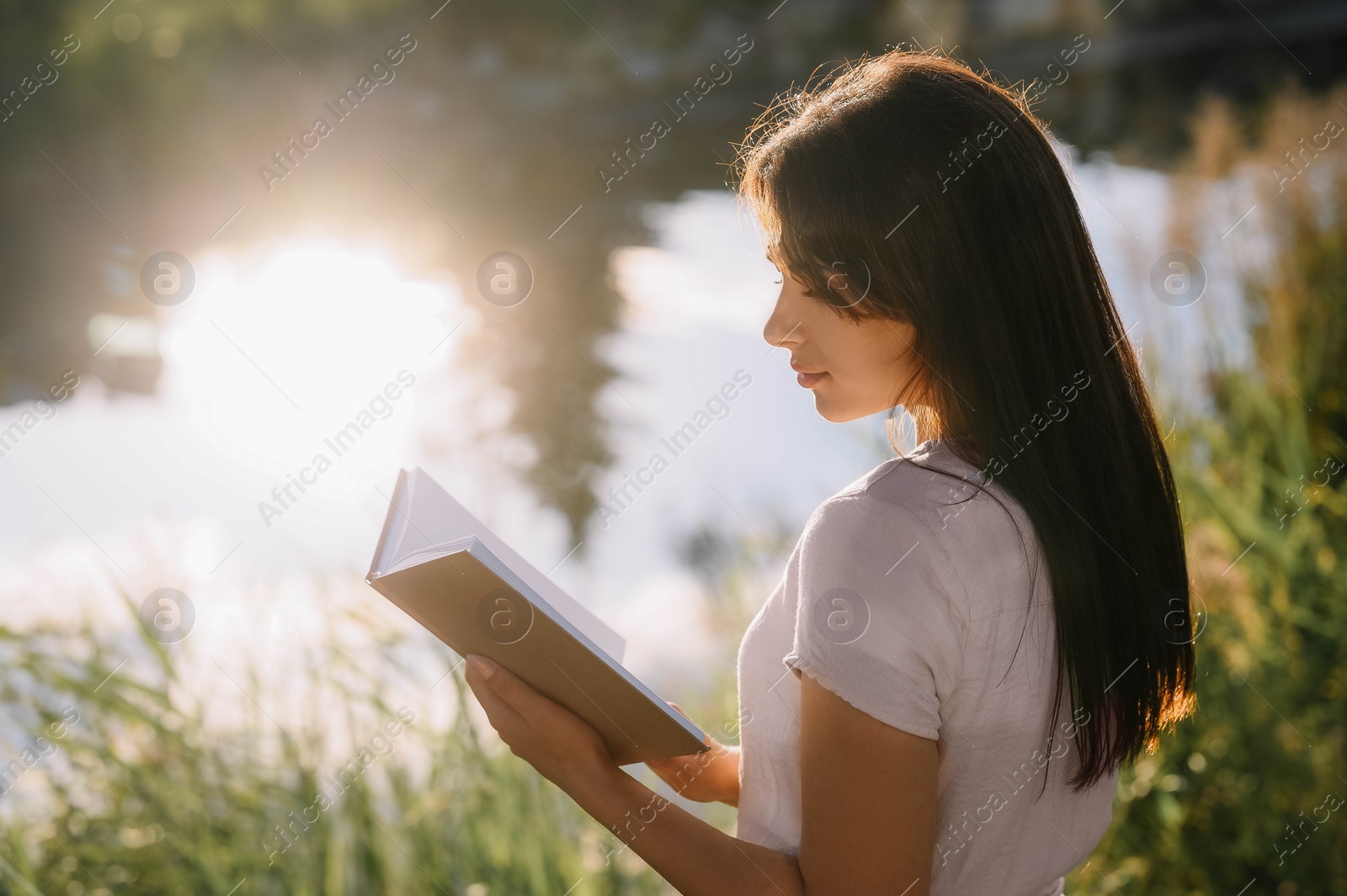 Photo of Young woman reading book near lake on sunny day