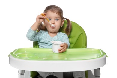Cute little child eating tasty yogurt from plastic cup with spoon in high chair on white background