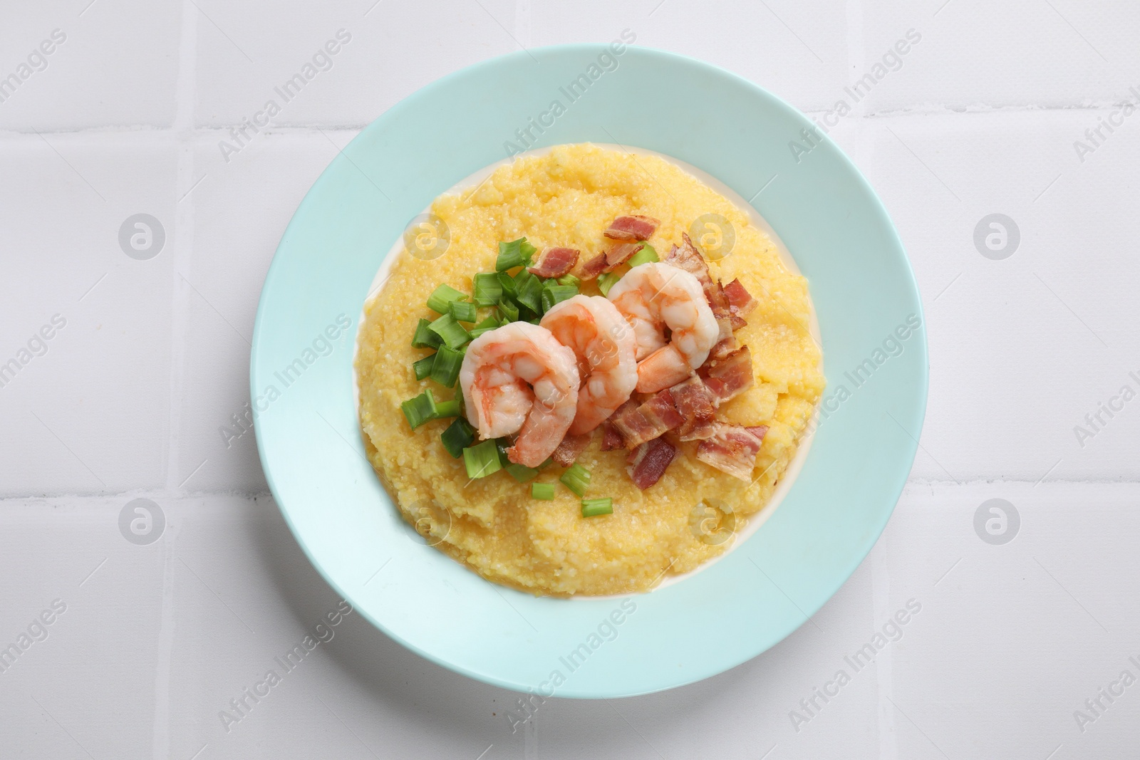 Photo of Plate with fresh tasty shrimps, bacon, grits and green onion on white tiled table, top view