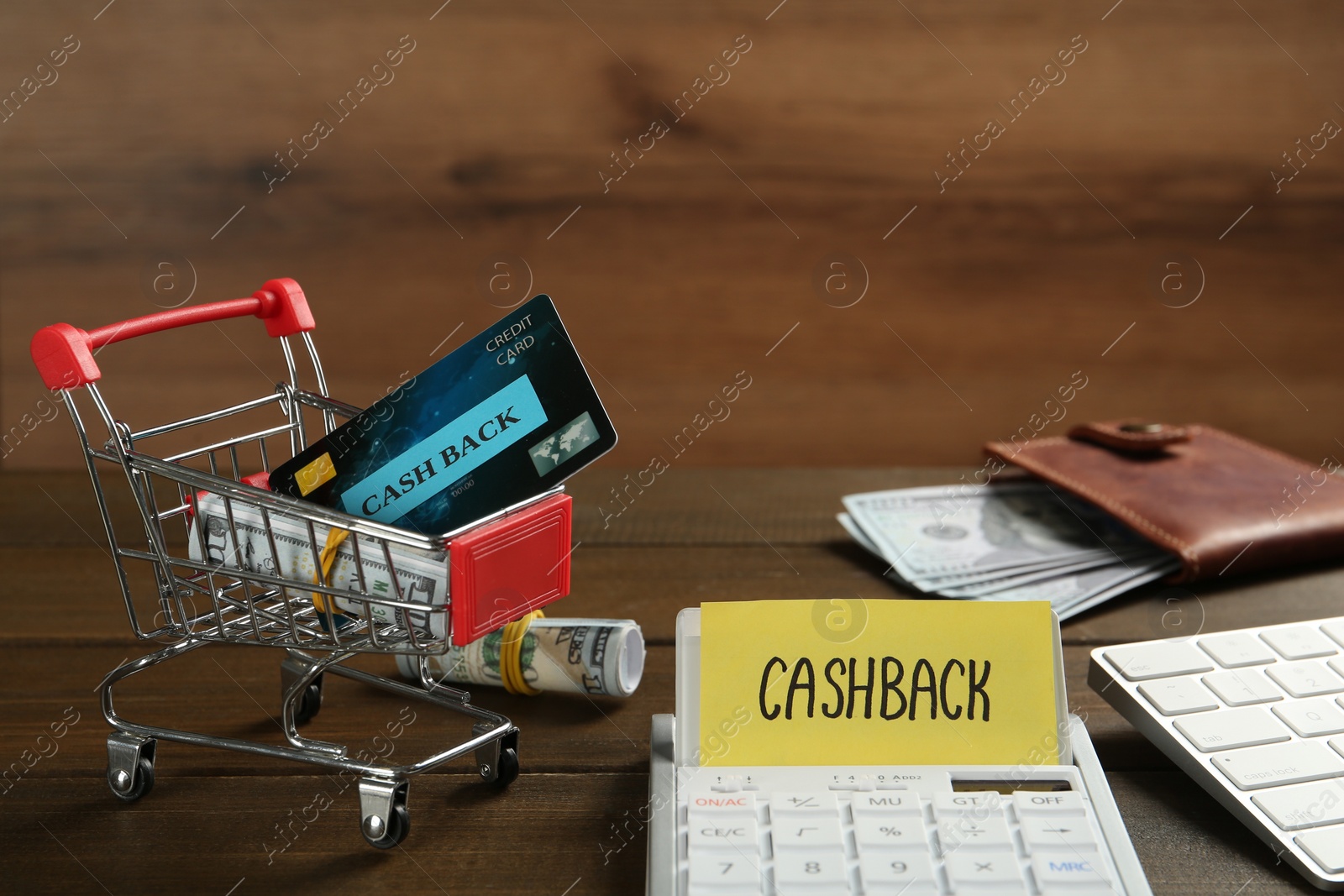 Photo of Calculator with sign Cashback near credit card and rolled banknotes in small shopping cart on wooden table. Space for text