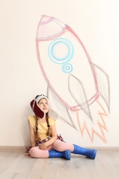 Adorable little child playing astronaut near wall with drawing of spaceship indoors