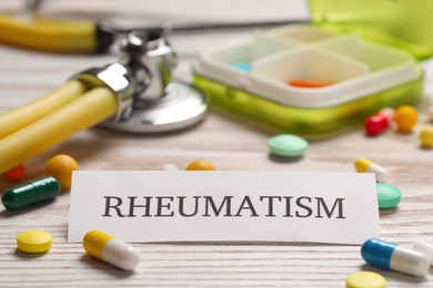 Card with word Rheumatism, stethoscope and pills on white wooden table, closeup