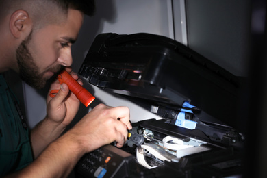Photo of Repairman with flashlight fixing modern printer indoors