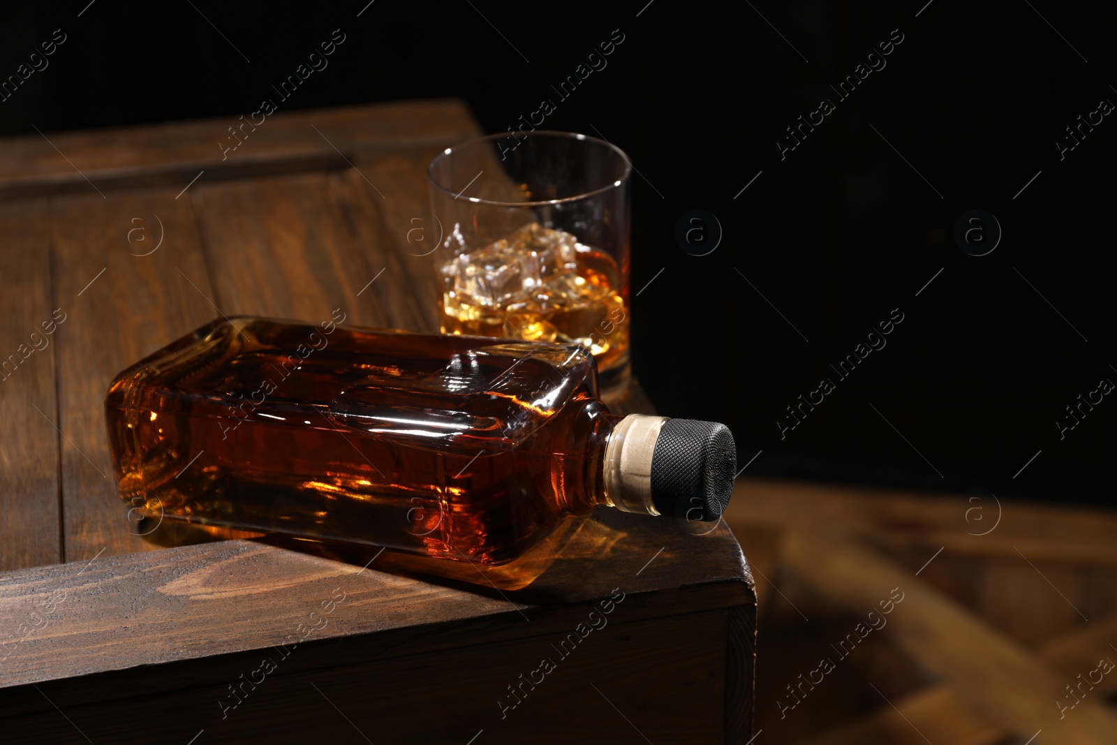 Photo of Whiskey with ice cubes in glass and bottle on wooden crate against black background, closeup. Space for text