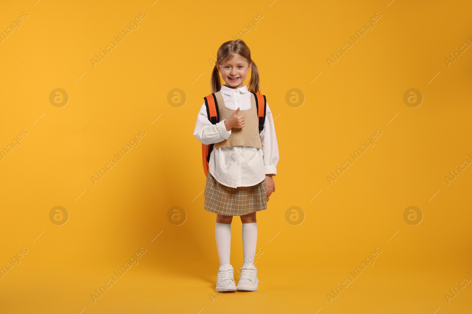 Photo of Happy schoolgirl with backpack showing thumb up gesture on orange background