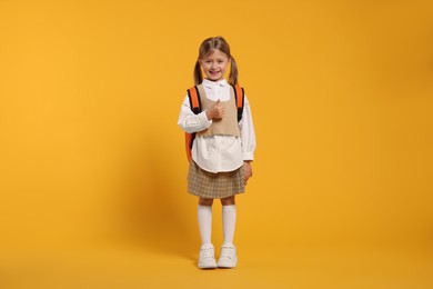 Happy schoolgirl with backpack showing thumb up gesture on orange background