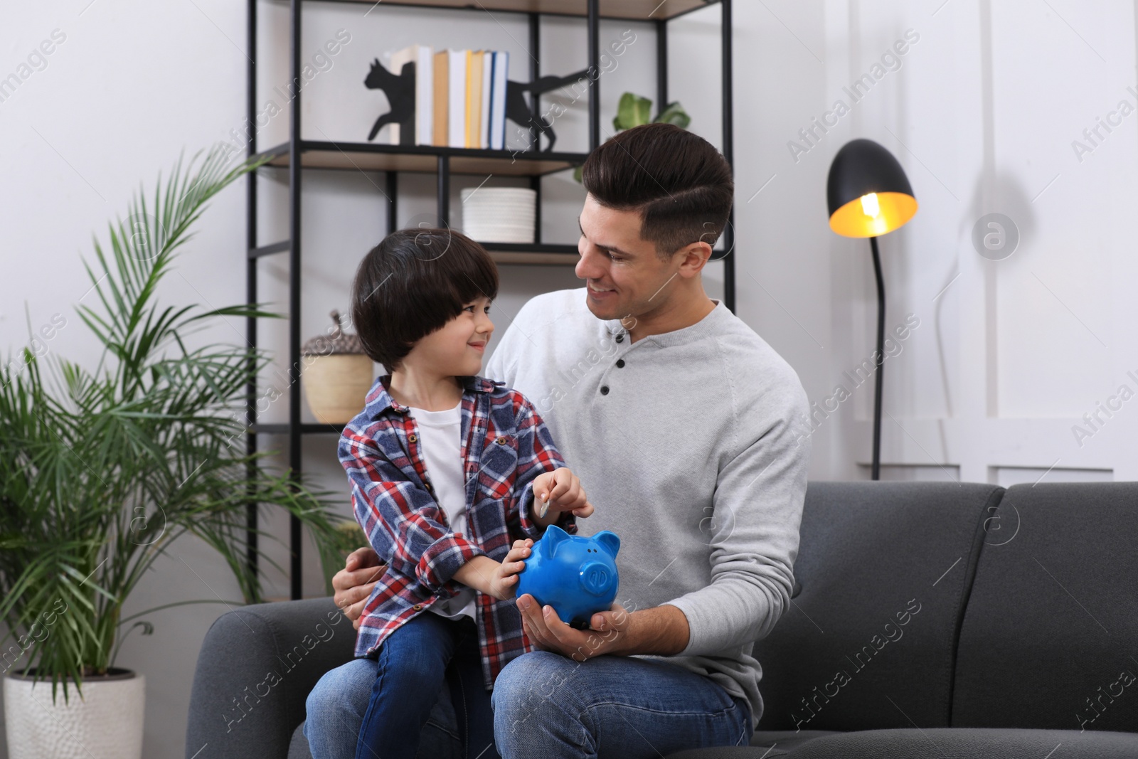 Photo of Little boy with his father putting coin into piggy bank at home