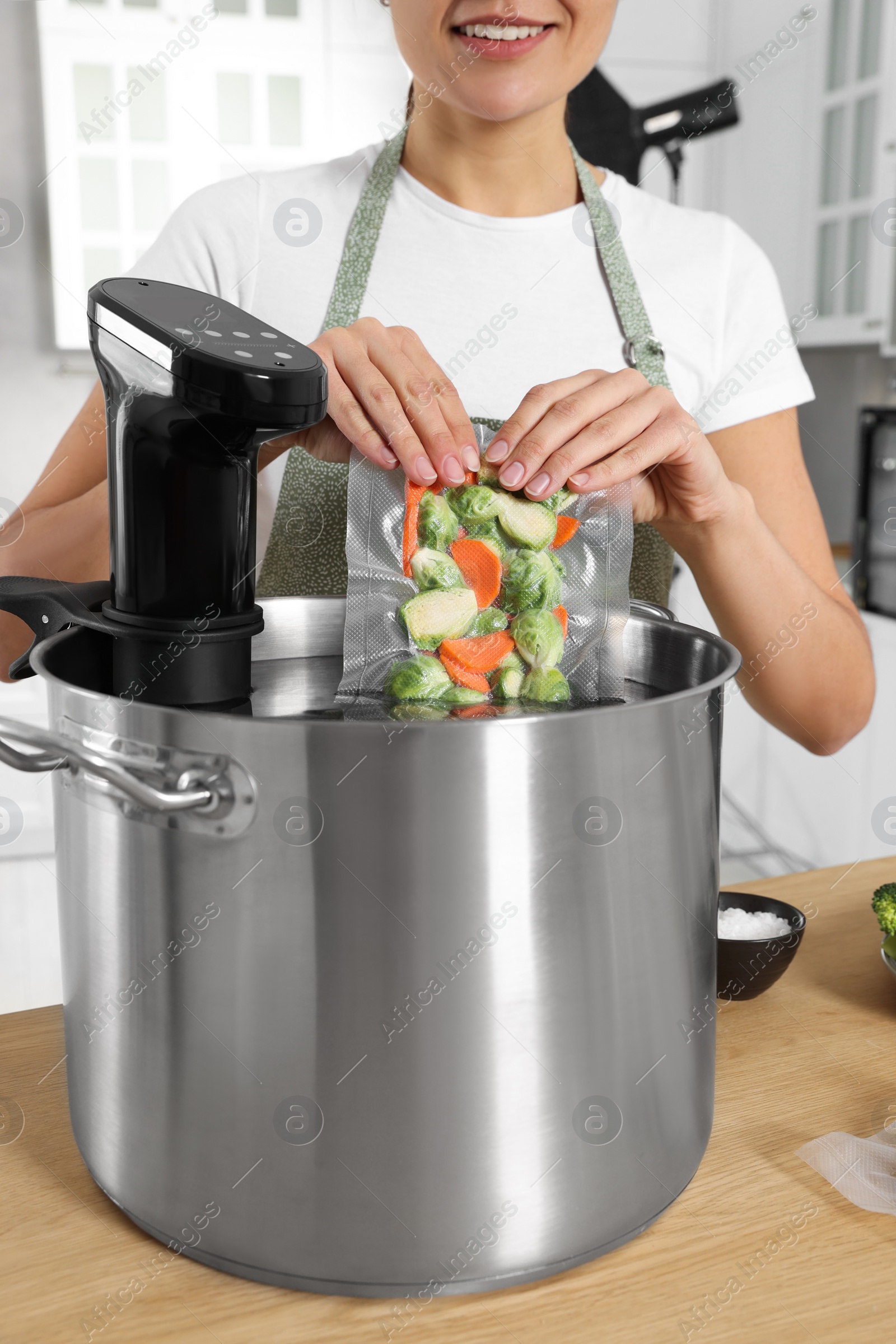 Photo of Woman putting vacuum packed vegetables into pot with sous vide cooker in kitchen, closeup. Thermal immersion circulator