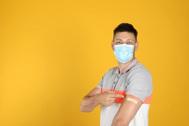 Vaccinated man with protective mask showing medical plaster on his arm against yellow background. Space for text