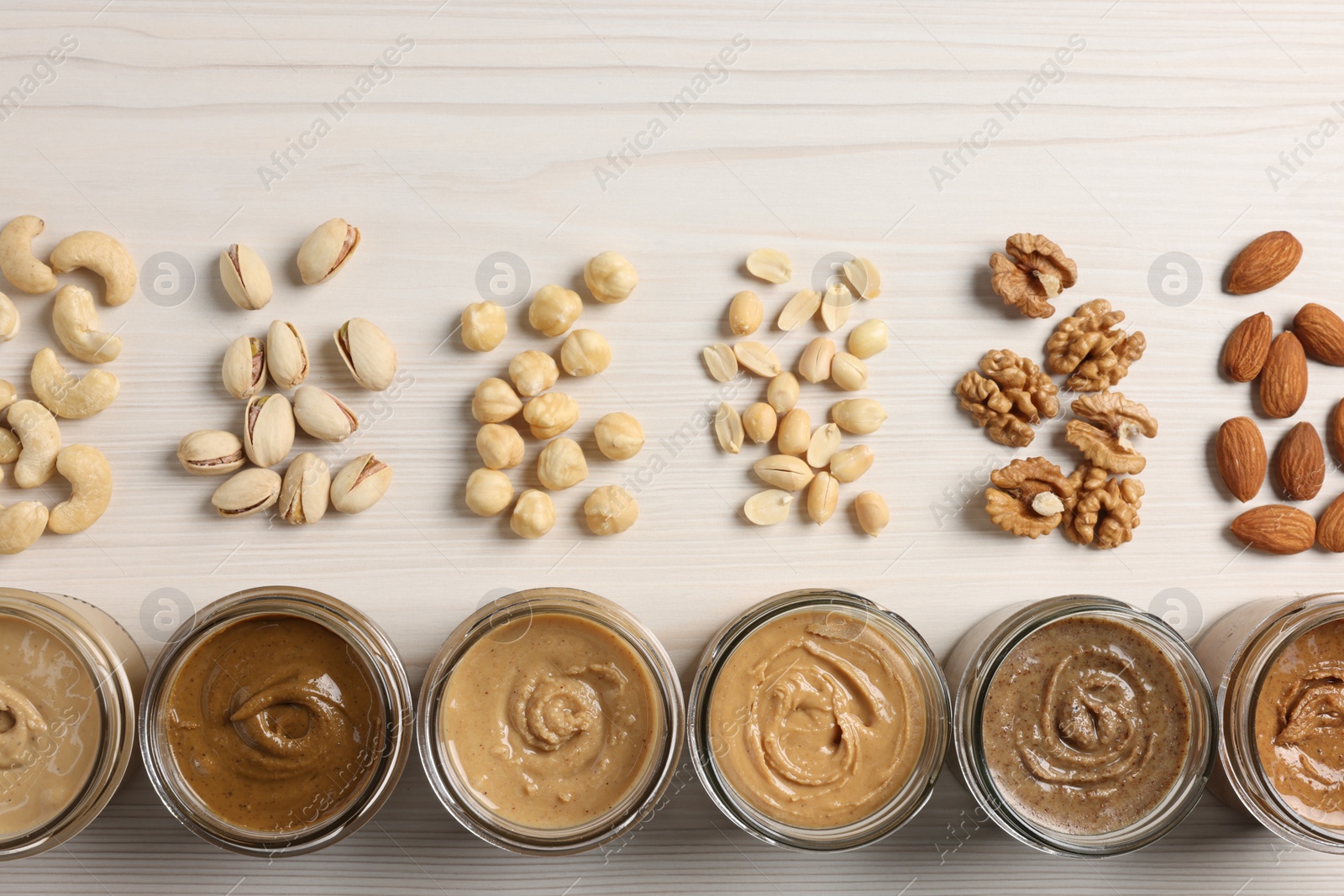 Photo of Many tasty nut butters in jars and nuts on white wooden table, flat lay