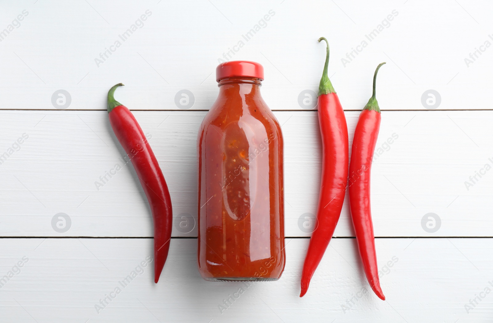 Photo of Spicy chili sauce in bottle and peppers on white wooden table, flat lay