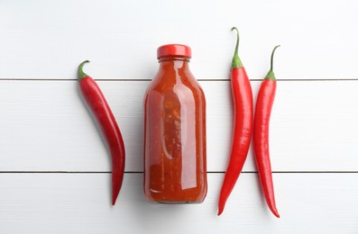 Photo of Spicy chili sauce in bottle and peppers on white wooden table, flat lay