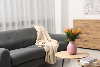 Photo of Vase with beautiful flowers and books on wooden nesting tables near grey sofa in room