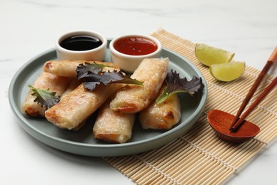 Photo of Tasty fried spring rolls, lettuce, lime and sauces on white marble table, closeup