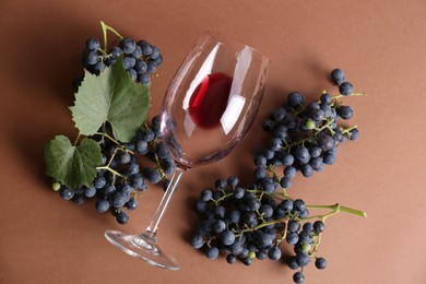 Photo of Overturned glass with red wine and grapes on brown background, flat lay