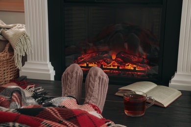 Woman with cup of tea and book resting near fireplace at home, closeup