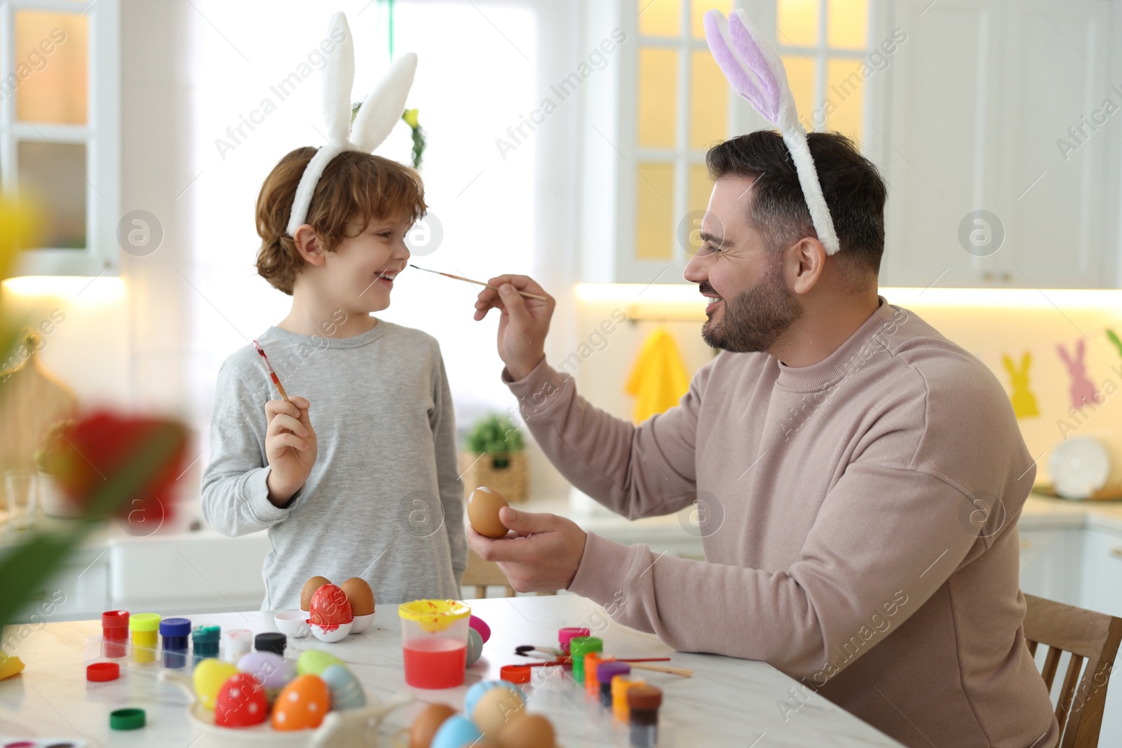 Photo of Easter celebration. Father with his little son having fun while painting eggs at white marble table in kitchen