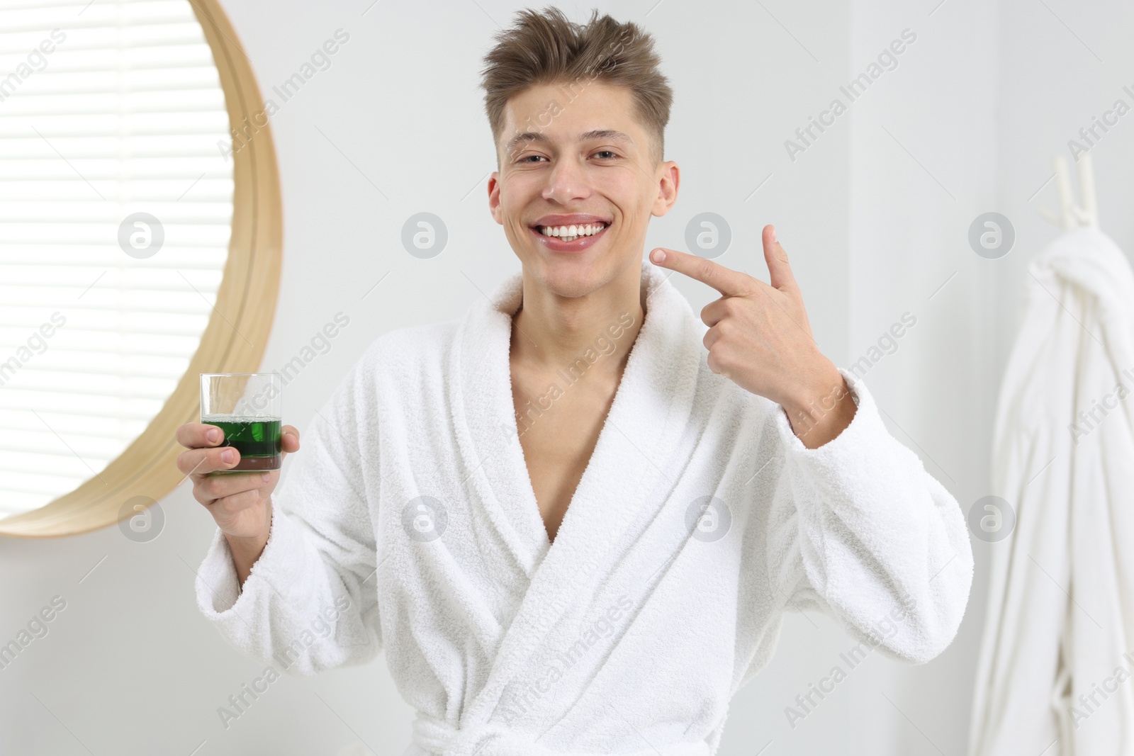 Photo of Young man with mouthwash pointing at his healthy teeth in bathroom