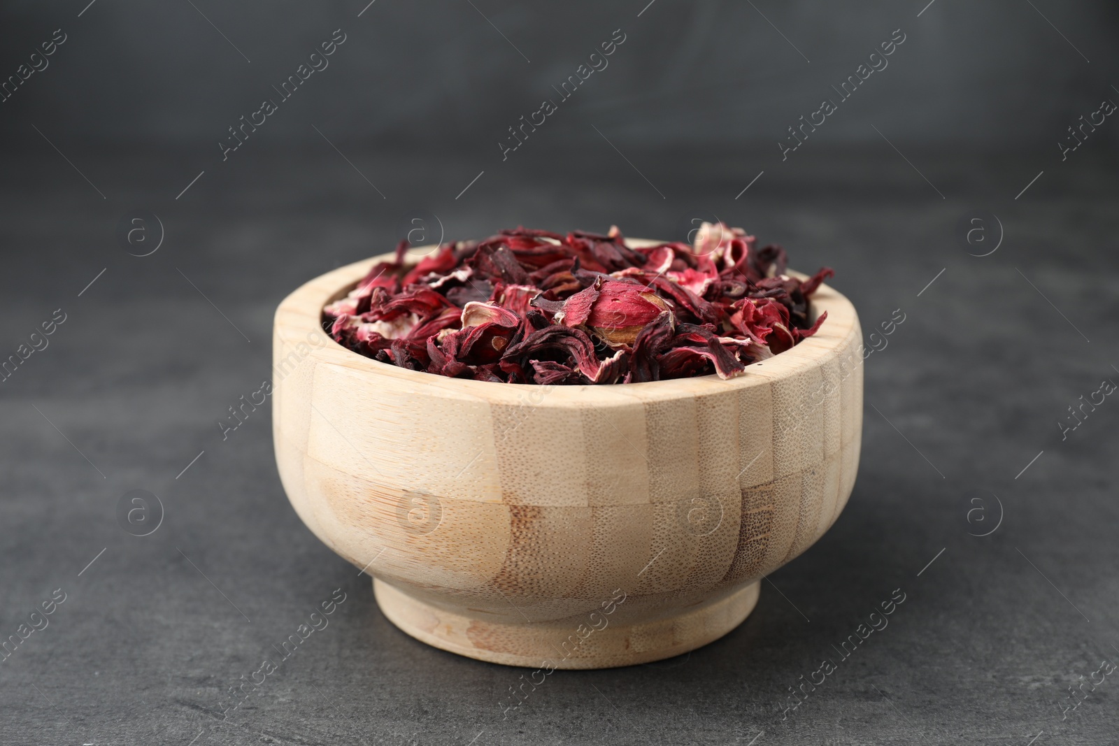 Photo of Hibiscus tea. Wooden bowl with dried roselle calyces on grey table, closeup