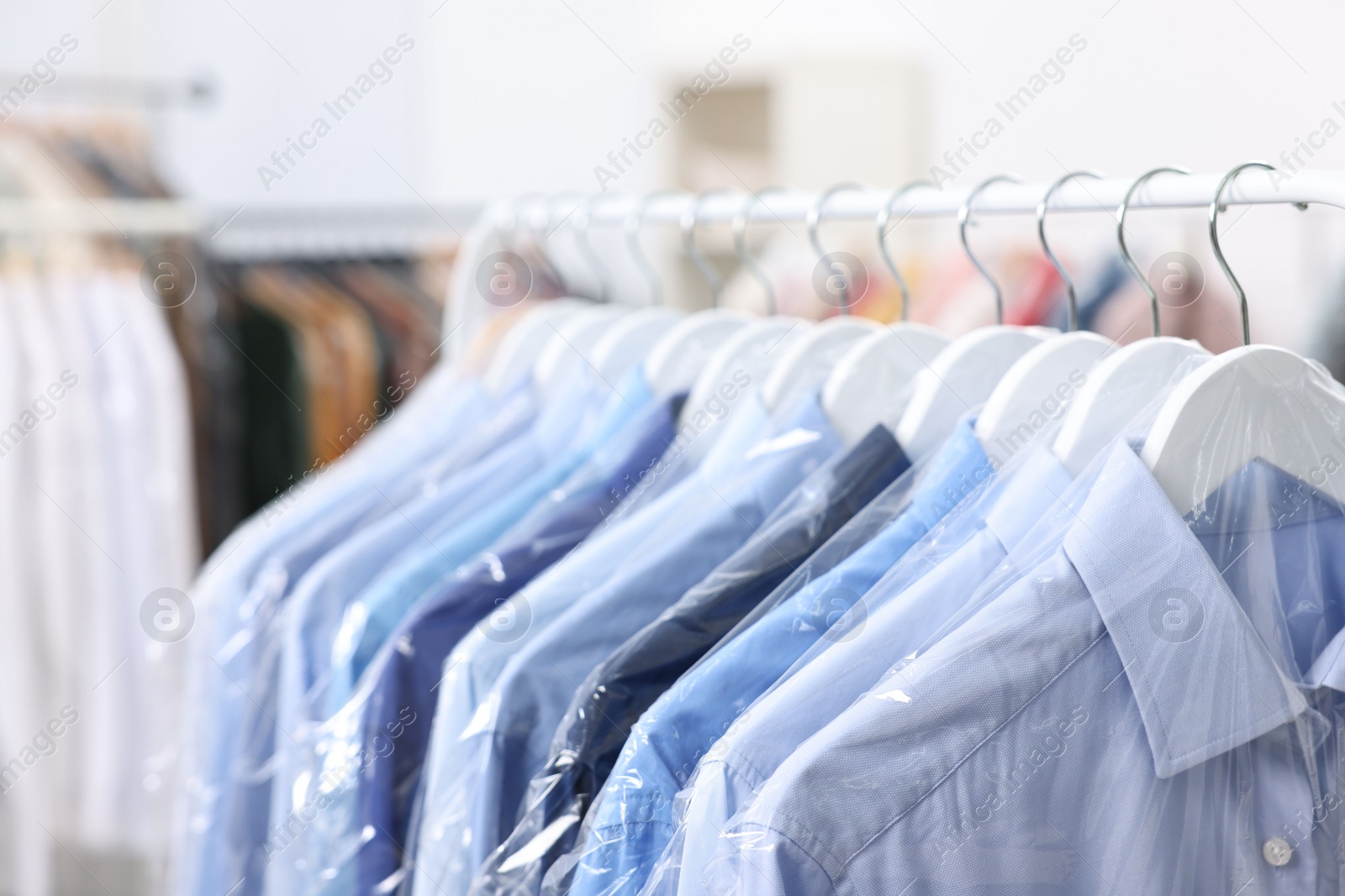 Photo of Dry-cleaning service. Many different clothes in plastic bags hanging on rack indoors, closeup