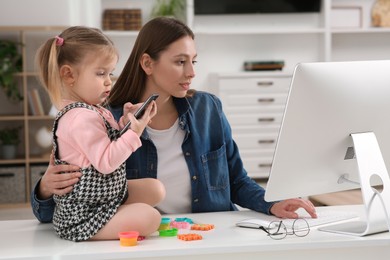 Woman working remotely at home. Mother using computer while her daughter playing with phone. Child sitting on desk