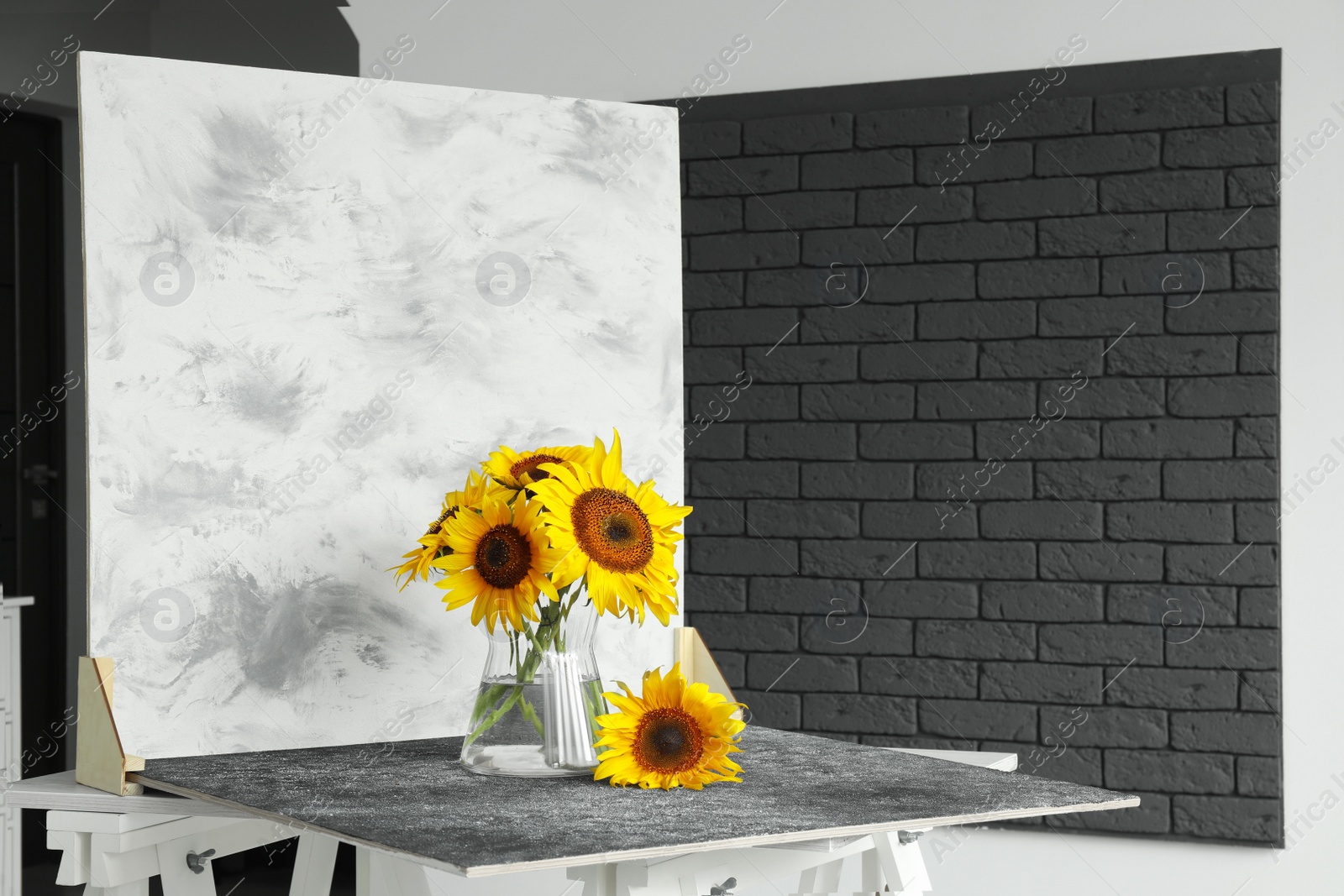 Photo of Glass vase with beautiful sunflowers and double-sided backdrop on table in photo studio