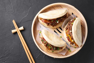 Photo of Delicious gua bao in bamboo steamer and chopsticks on black table, top view