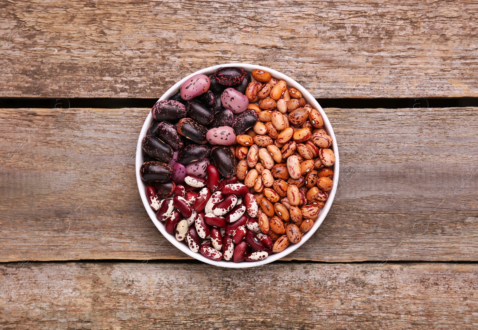 Photo of Different kinds of dry kidney beans in bowl on wooden table, top view