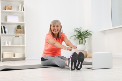 Photo of Senior woman in sportswear stretching near laptop on mat at home