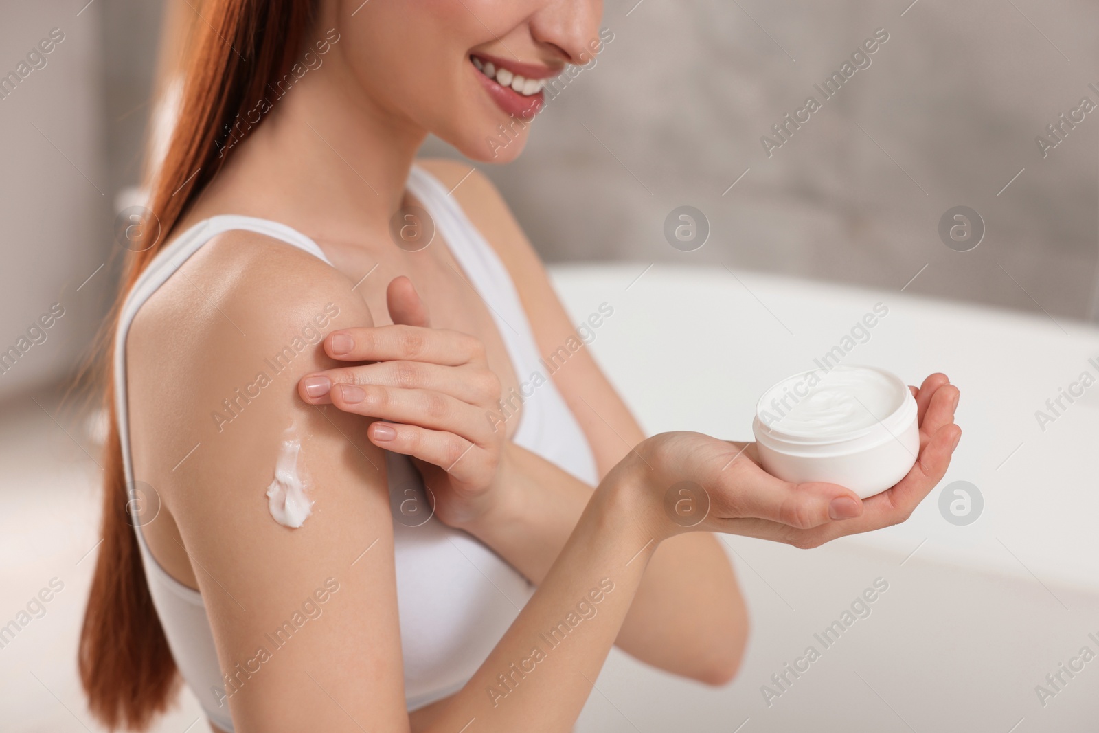 Photo of Young woman applying body cream onto shoulder in bathroom, closeup