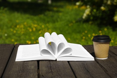 Photo of Open book and paper coffee cup on wooden table outdoors. Space for text