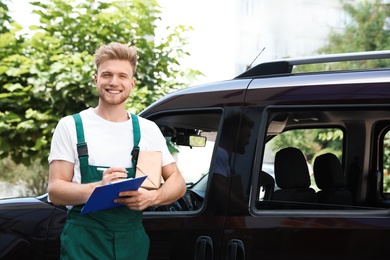 Photo of Young courier with clipboard and parcel near delivery car outdoors