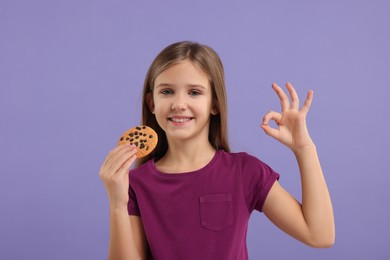 Photo of Cute girl with chocolate chip cookie showing OK gesture on purple background