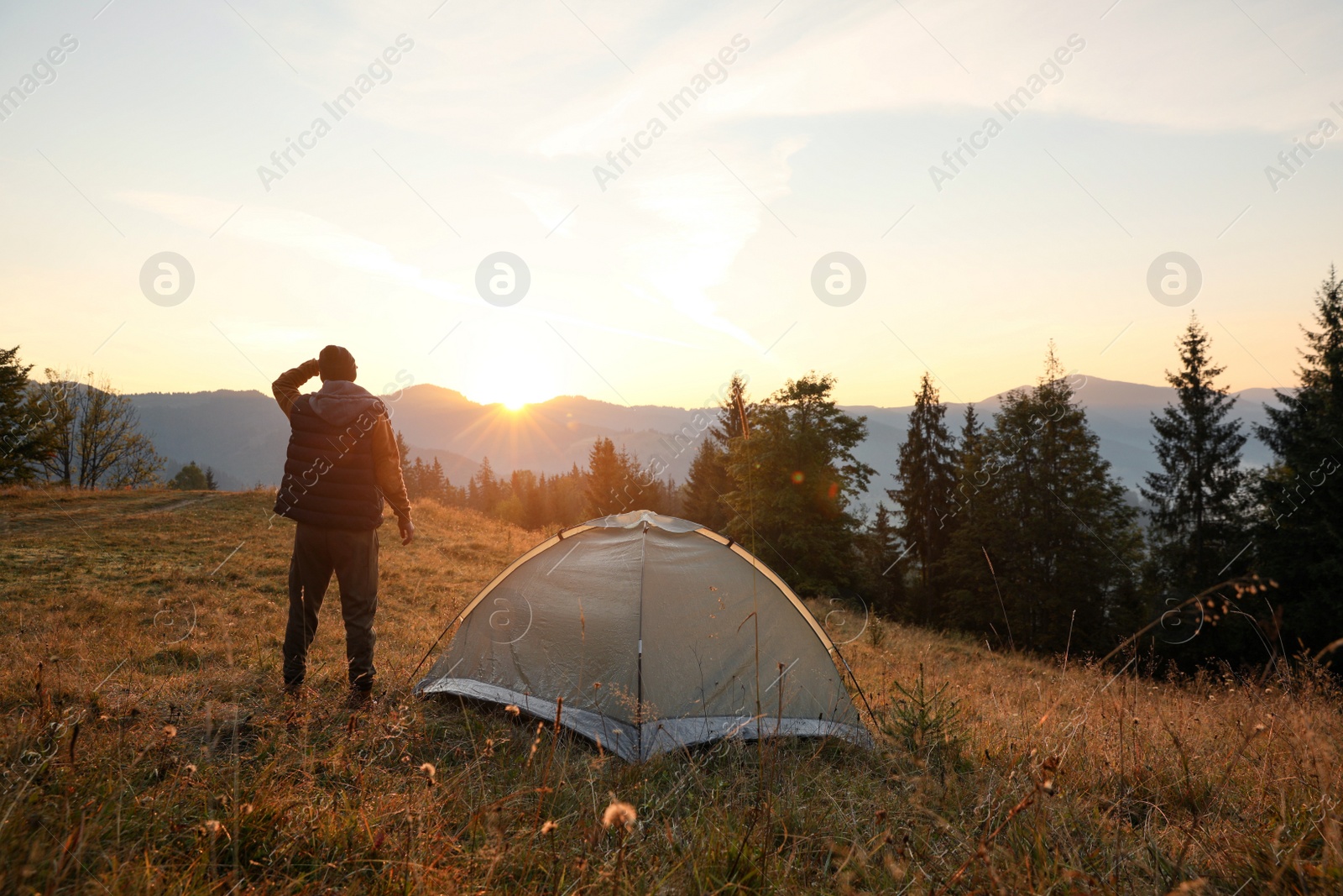 Photo of Man near camping tent in mountains at sunset, back view